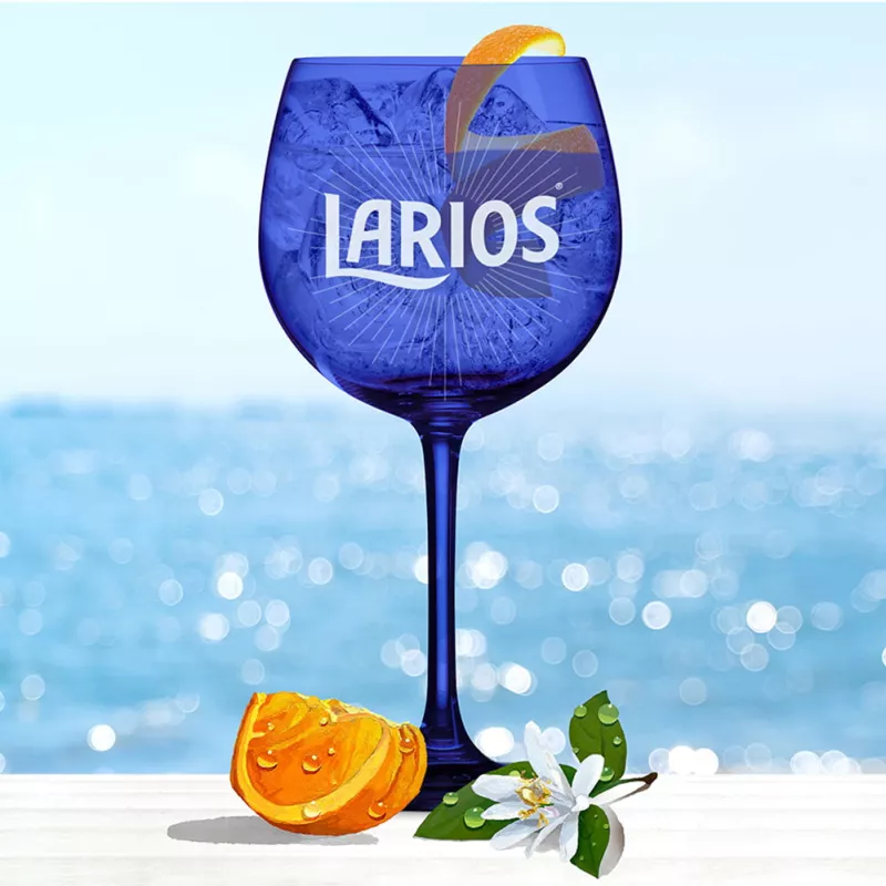 Blue Larios glass cup filled with Gin Tonic Rose and garnished with orange peels, standing on a white surface infront of the ocean