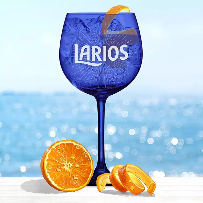 Blue Larios glass cup filled with Gin Rose Lemonade and garnished with orange peels, standing on a white surface infront of the ocean