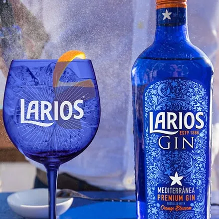 A person spritzing water at a blue Larios glass cup and a bottle of Larios Gin on a blue surface
