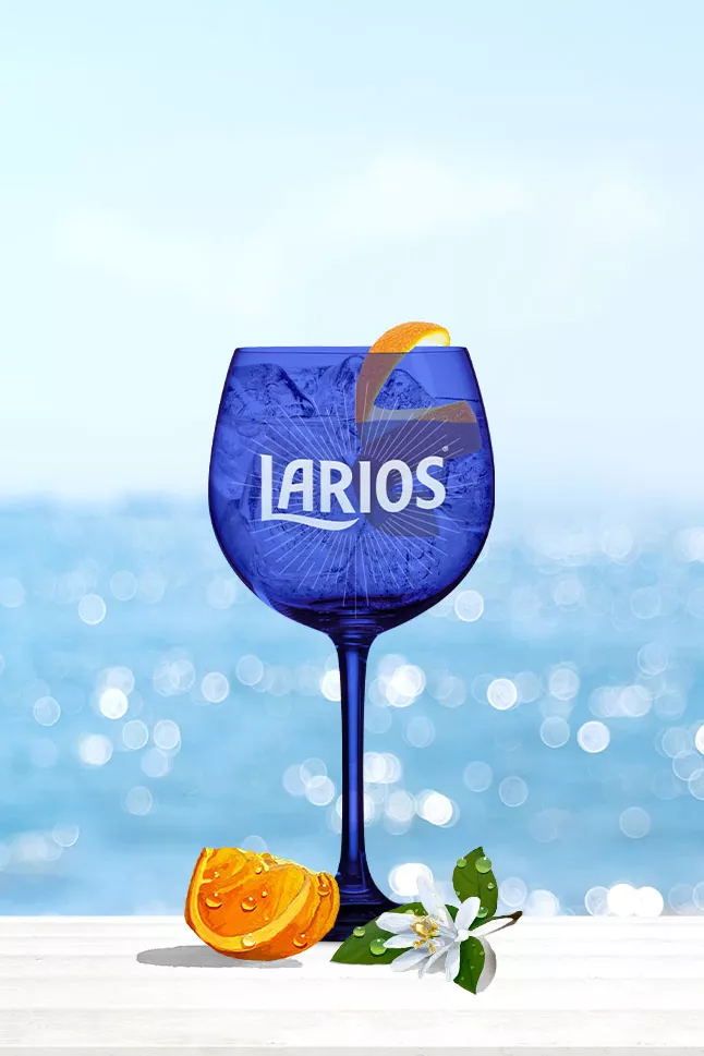 Blue Larios glass cup filled with Gin Tonic Rose and garnished with orange peels, standing on a white surface infront of the ocean
