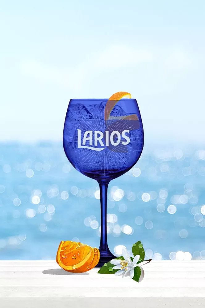 Blue Larios glass cup filled with Gin Tonic Rose and garnished with orange peels, standing on a white surface infront of the ocean.