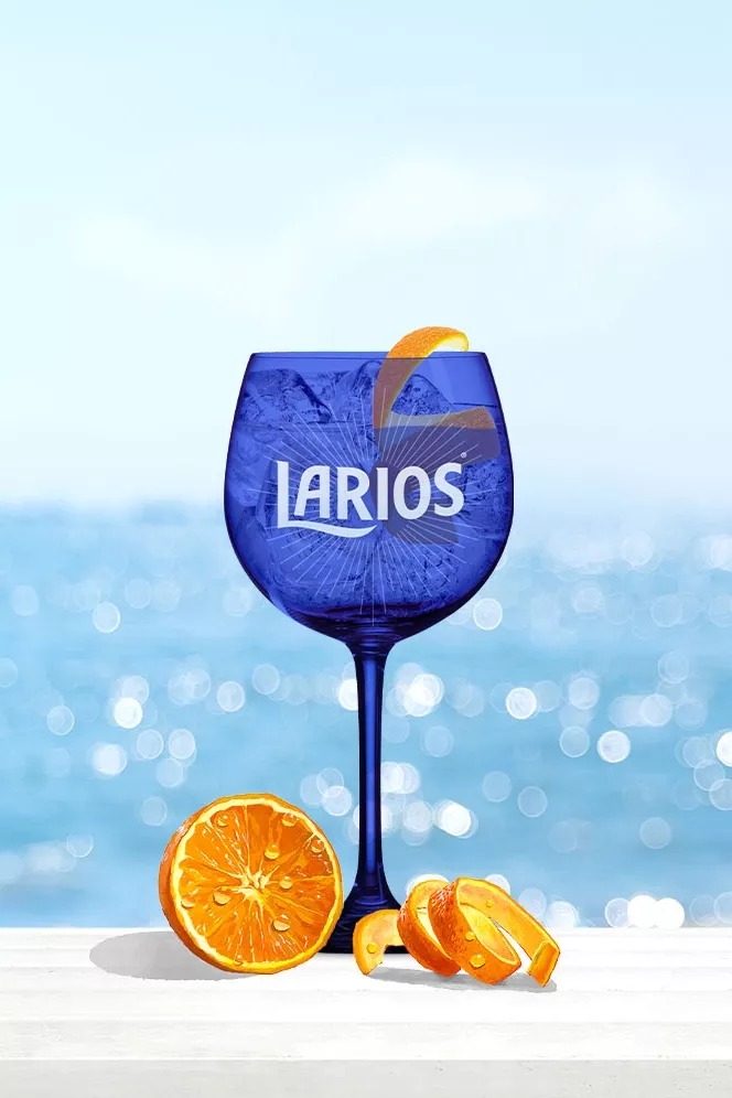 A glass of Larios Gin Rose Lemonade in front of a beach background with orange piece.