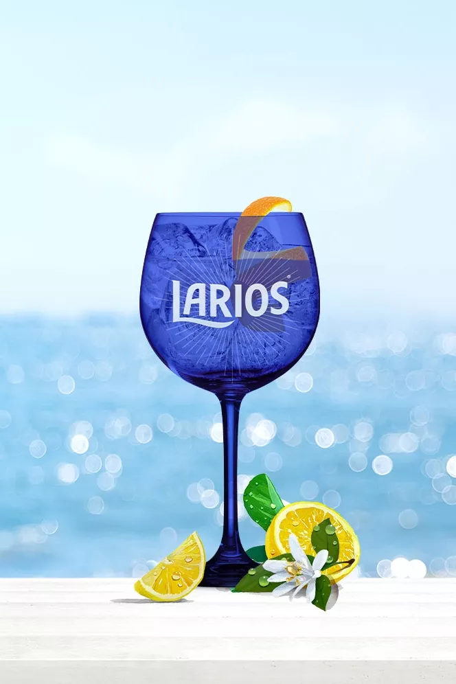 A glass of Larios Gin Tonic Mediterranean cocktail with lemon pieces in front of a beach background.