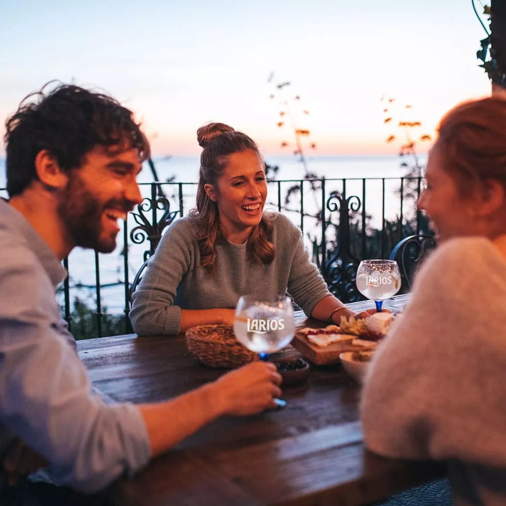 (Gallery_2) Alt: Two late-twenties women and a man enjoy conversation over gin cocktails and appetizers, relaxing at an old-world wooden table at a sea-side cafe. | Filename: friends-toast-gin-cocktails.jpg