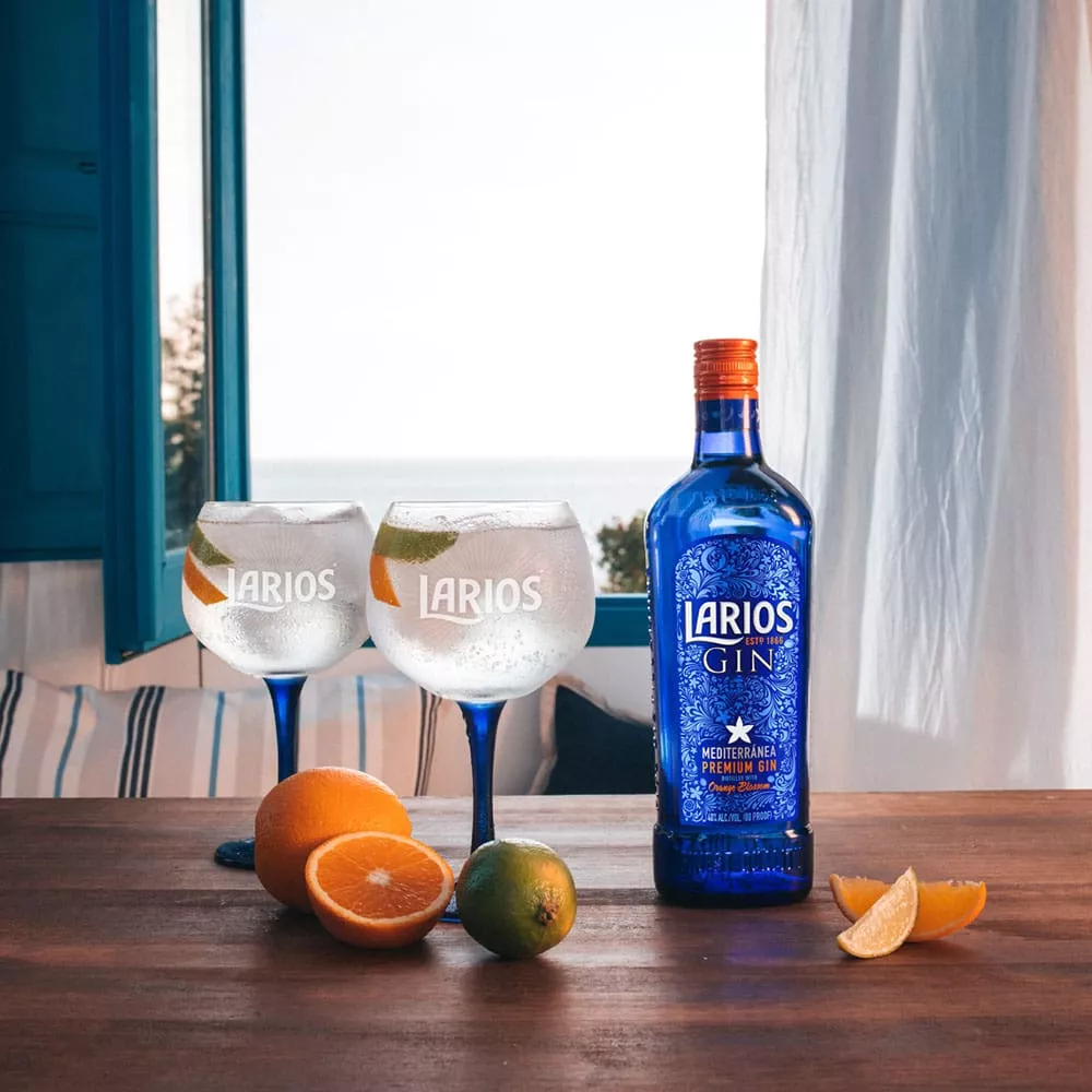 Two Larios glasses and a bottle of Larios Gin standing on a wooden surface in a room with orange and limes