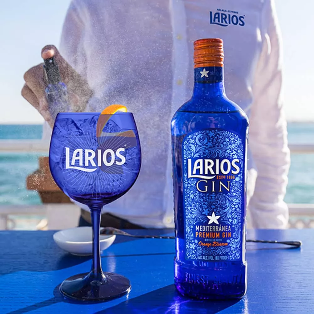 A person spritzing water at a blue Larios glass cup and a bottle of Larios Gin on a blue surface