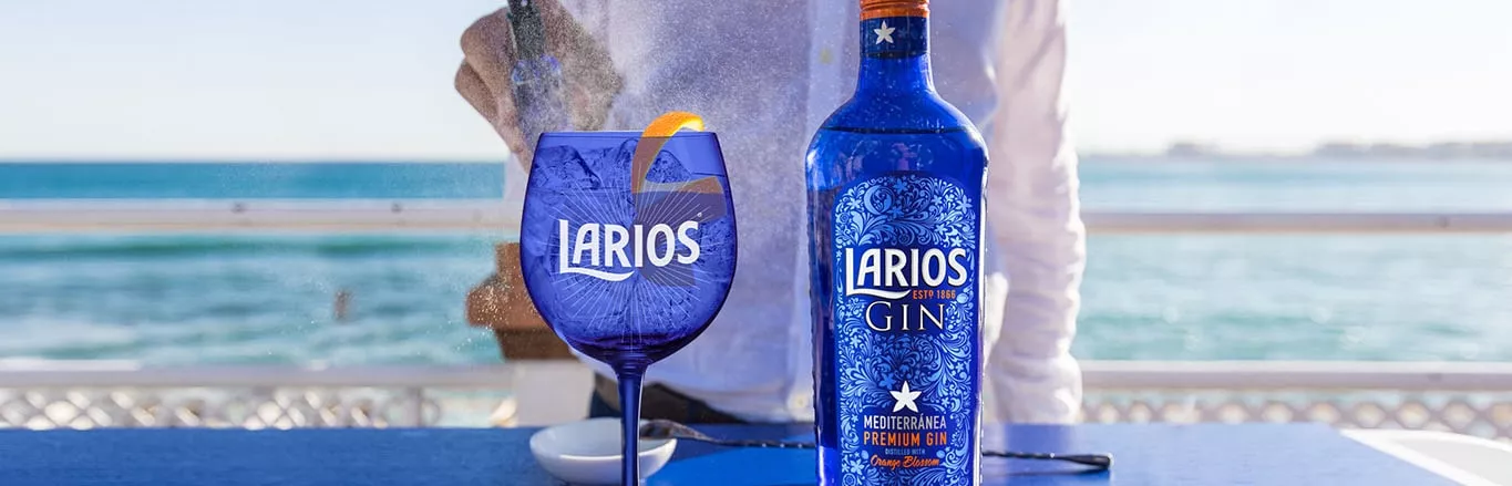 A person spritzing water at a blue Larios glass cup and a bottle of Larios Gin on a blue surface